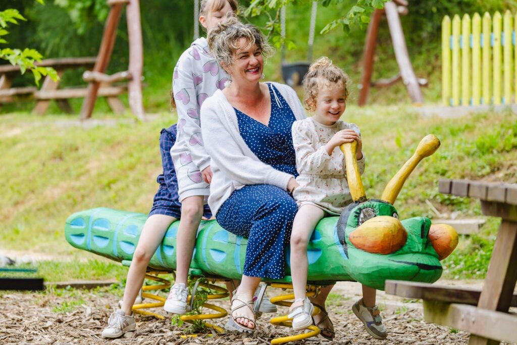 Une mère partage un moment complice avec ses deux filles sur un jeu à ressort en forme de chenille verte. L'éclat souriant de toute la famille reflète une ambiance chaleureuse et amusante.
