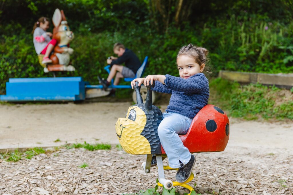 Une petite fille s'amuse sur un jeu à ressort en forme de coccinelle rouge et noire. L'arrière-plan montre d'autres enfants jouant sur des structures similaires, créant une atmosphère conviviale.