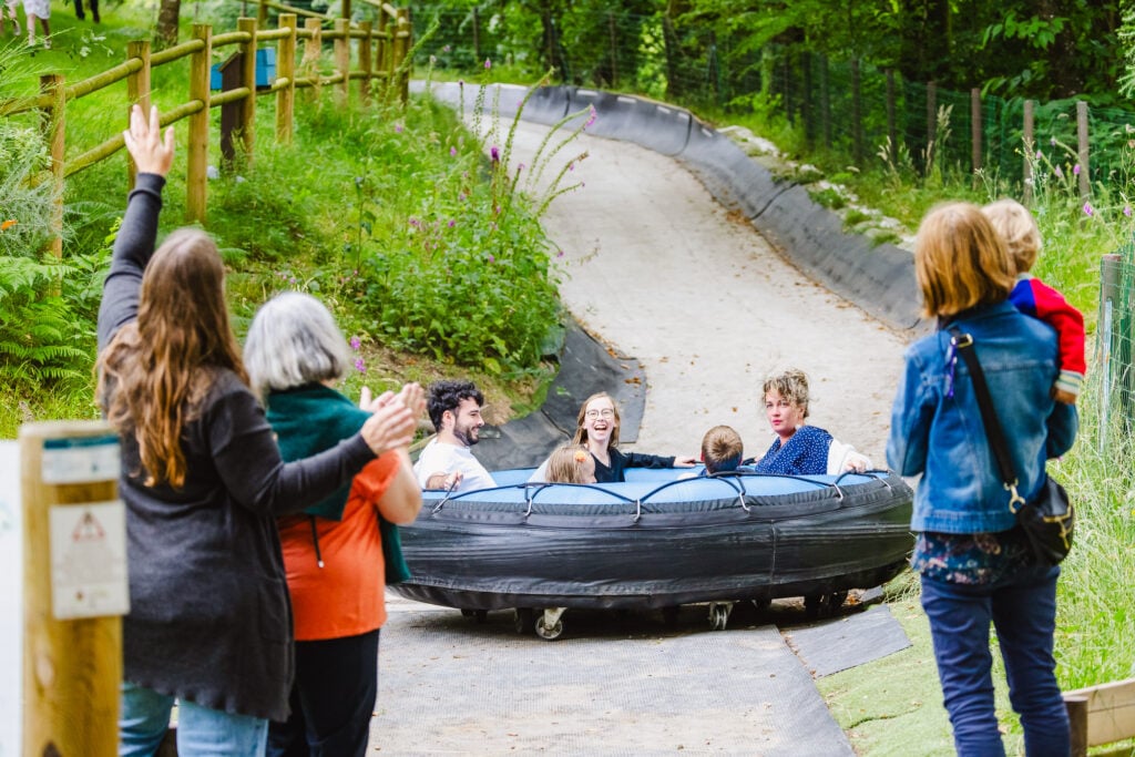 Une scène animée de loisirs en plein air capturée sur une piste de luge d'été à La Balade du Père Nicolas. Des visiteurs, deux adultes et deux enfants, rient et crient de joie tout en glissant sur une grande bouée noire sur la piste courbée. Leurs expressions joyeuses sont accueillies par les applaudissements et les encouragements des spectateurs qui se respectent le long du parcours. Les bords de la piste sont bordés de végétation dense et de fleurs sauvages, ajoutant une touche de nature au décor de cette activité de plein air. air."