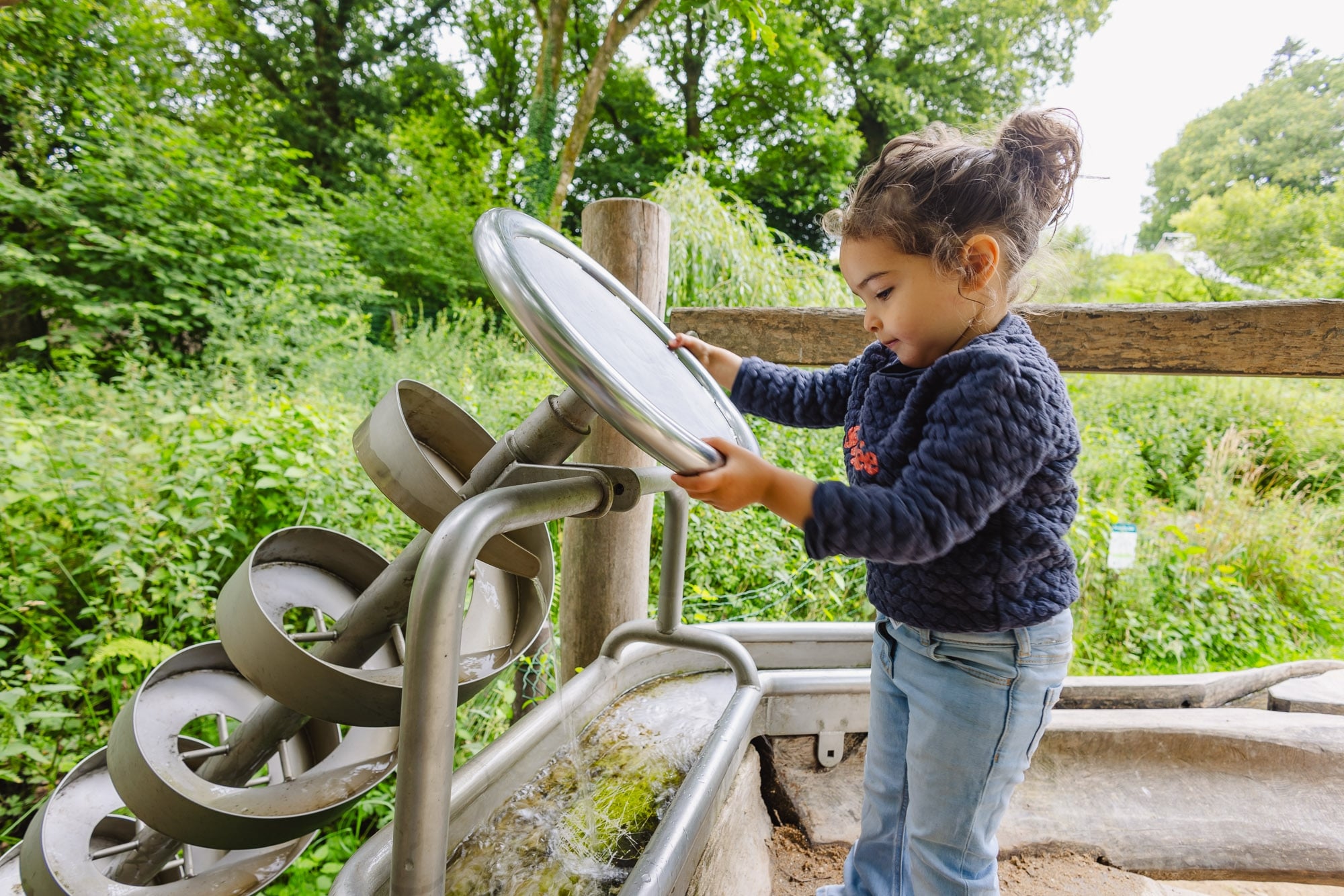 Une petite fille concentrée tourne une roue métallique pour faire remonter de l'eau dans un mécanisme interactif. Le cadre est entouré d'une végétation luxuriante, ajoutant une atmosphère naturelle et paisible.