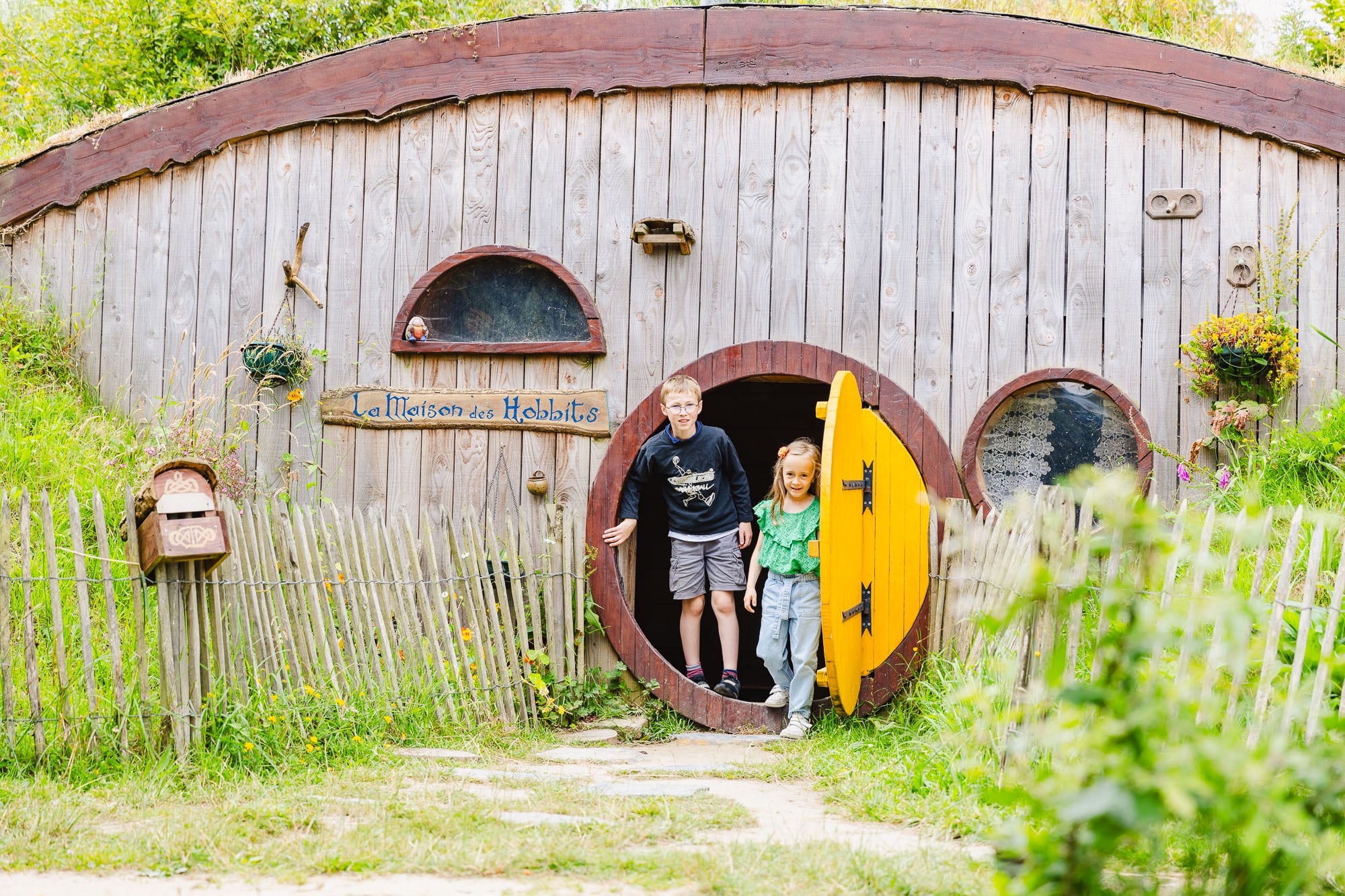 Deux enfants sortent joyeusement par la porte ronde jaune de la Maison des Hobbits, un lieu enchanteur niché dans la verdure avec une clôture en bois et des détails charmants.