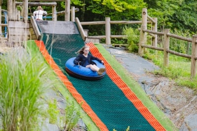 Une jeune fille glissant avec enthousiasme sur une piste de tubing, installée sur une bouée, dans un cadre naturel bordé de verdure et de barrières en bois.