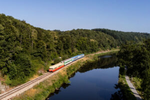 Cette photo montre le train Napoléon Express serpenter à travers une campagne paisible. Le train, peint en rouge et blanc, suit une voie ferrée qui longe un plan d'eau calme, reflétant le paysage verdoyant des arbres denses sur les collines environnantes. C'est une journée ensoleillée avec un ciel clair, ce qui accentue les couleurs vives de la nature et du train lui-même.