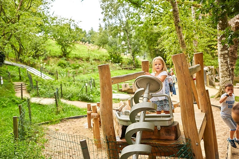 Une jeune fille enthousiaste tourne une roue pour activer un jeu d'eau composé de spirales en métal. L'installation est située dans un cadre verdoyant avec un chemin sinueux à l'arrière-plan. Son expression joyeuse et l'activité environnante donnent vie à la scène, soulignant l'interaction ludique avec l'eau.
