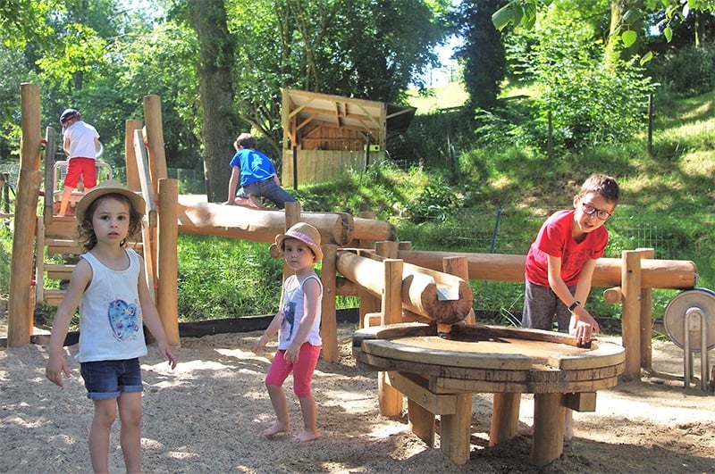 Des enfants de divers âges s'amusent autour d'un jeu d'eau en bois dans un parc. Un garçon actif tourne une manivelle en bois pour manipuler l'eau tandis que deux petites filles observent attentivement. Le jeu est situé dans une aire de sable, sous un ciel ensoleillé, ce qui crée une atmosphère joyeuse et vivante.
