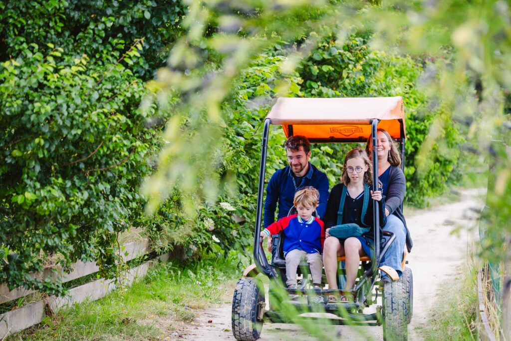 Une famille de quatre personnes, dont un enfant en bas âge, fait une balade en kart à quatre places couvert par un toit orange. Ils profitent d'une journée fraîche, entourés de végétation dense et luxuriante.