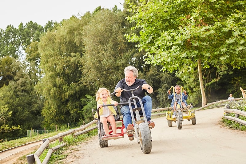 Un grand-père et sa petite-fille, suivis par d'autres enfants, rient tout en faisant la course sur des karts à pédales sur un chemin sinueux entouré de verdure luxuriante.