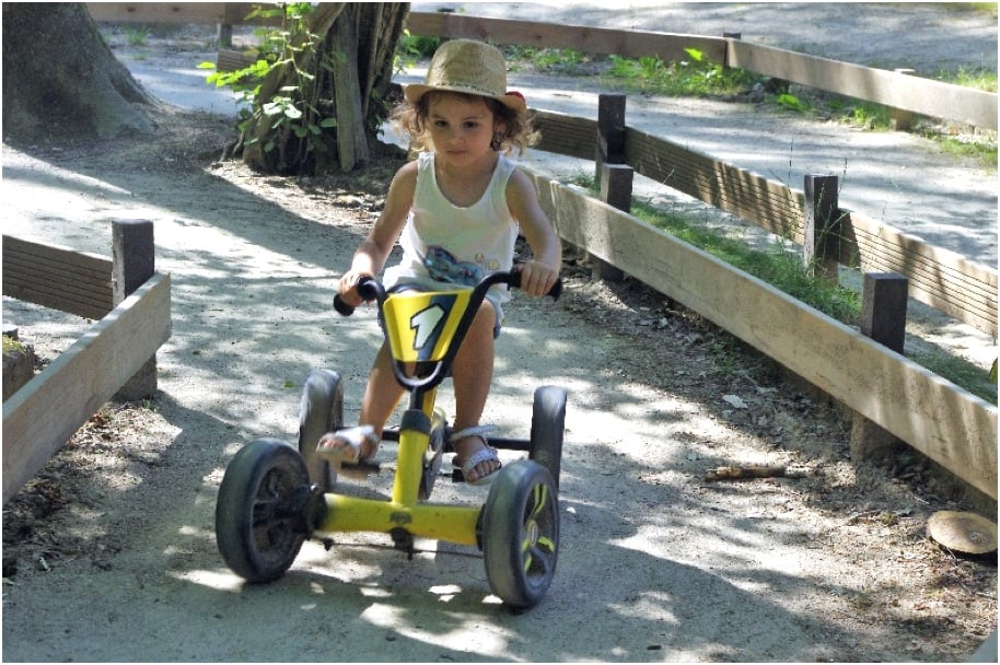 Une petite fille avec un chapeau de paille conduit un kart à pédales jaune le long d'un chemin bordé de bois. Elle affiche un air déterminé et curieux, explorant le parc en toute autonomie.