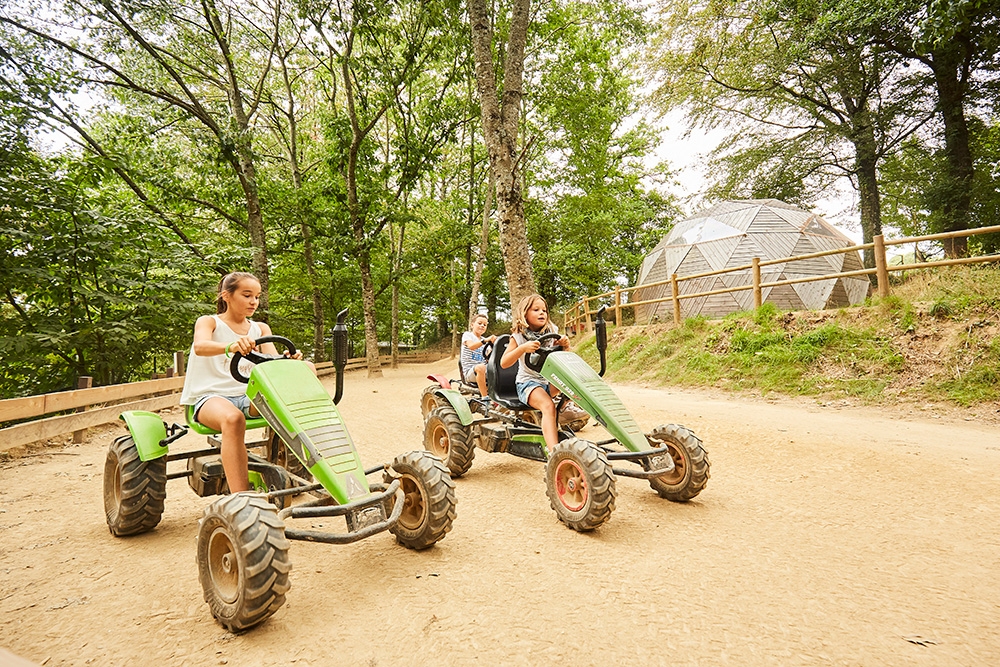 Trois enfants pilotent des karts à pédales sur un chemin de terre entouré d'arbres dans un parc. Ils semblent se concentrer intensément sur leur course, le tout sous un ciel légèrement nuageux.