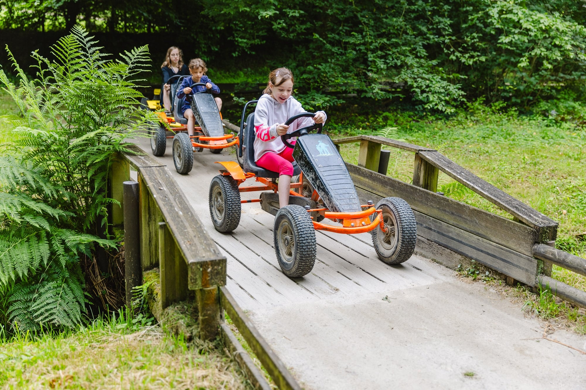 Trois enfants pilotent des karts à pédales sur un chemin de terre entouré d'arbres dans un parc. Ils semblent se concentrer intensément sur leur course, le tout sous un ciel légèrement nuageux.