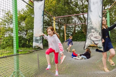 Un groupe d'enfants s'amuse dans un filet suspendu, courant et sautant joyeusement. Une jeune fille traverse la zone en tenant des coussins suspendus, ajoutant une touche d'aventure à cette aire de jeu unique en pleine nature.